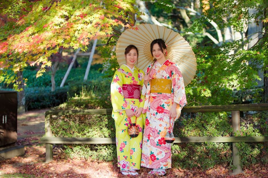 Two women in colorful kimonos with an umbrella posing in a serene Japanese garden setting.