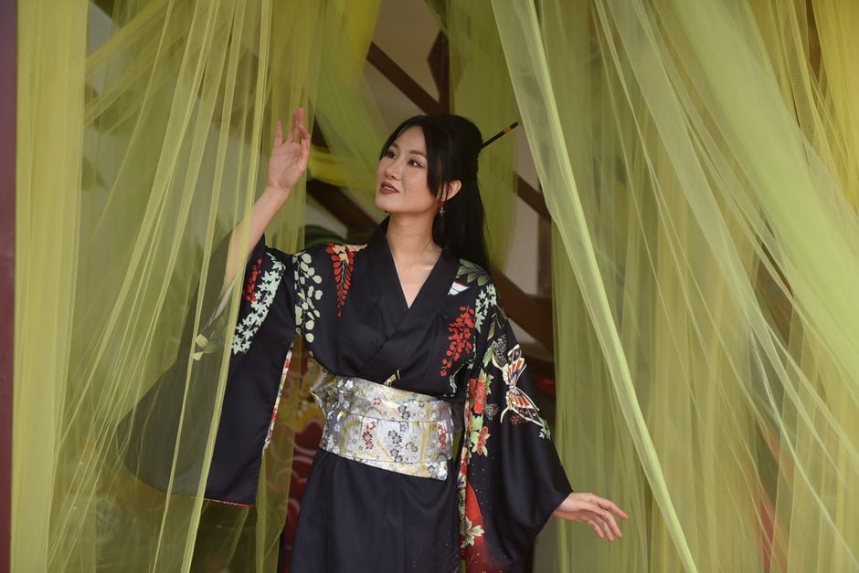A woman in a traditional floral kimono gracefully stands among sheer curtains.